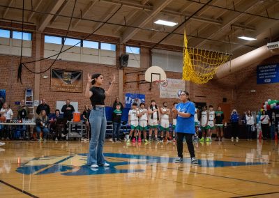 A photograph of two individuals standing in the center of the ASDB gymnasium. They are signing the National Anthem in ASL. Behind them, multiple individuals, including the Washington Terrier boys team, stand with their hands over their hearts.