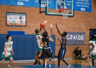 A photograph of an active WSBC basketball game in the ASDB gymnasium. On court are the competing Phoenix Roadrunner and Washington Terrier boys teams. In the photo, a Terrier player is attempting to drive through three Roadrunner defenders for a layup attempt.