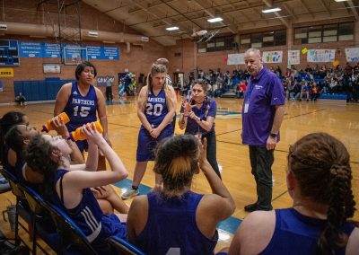 A photograph of an active WSBC basketball game in the ASDB gymnasium. On court are the competing Oregon Panther and Washington Terrier girls teams. In the photo, the Oregon Panther coach energetically signs to her players during a timeout break.