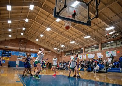 A photograph of an active WSBC basketball game in the ASDB gymnasium. On court are the competing Oregon Panther and Washington Terrier girls teams. In the photo, both teams react as a shot attempt sinks right through the bottom of the net.