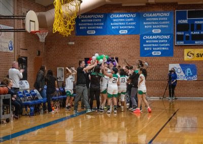 A photograph of an active WSBC basketball game in the ASDB gymnasium. On the court is the entire Washington Terrier girls team. They are gathered in a large circle and celebrating with their coaching staff.