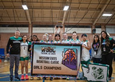A photo of the entire Washington Terrier girls team in the ASDB gymnasium. They are posing together with large smiles on their faces. They are collectively holding up a large banner reading, 'WSBC Tucson, AZ 2024 Western States Basketball Classic Girls Champions'.