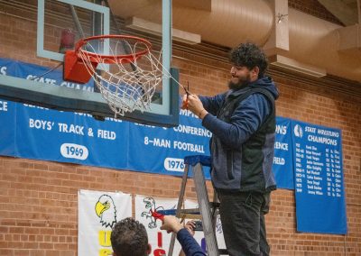 A photo of the Phoenix Roadrunner's assistant coach in the ASDB gymnasium. After winning the championship with his team, he is standing at the top of a ladder, cutting off a piece of the basketball net.