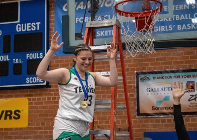 A photo of a Washington Terrier girls player in the ASDB gymnasium. After winning the championship with her team, she is standing at the top of a ladder, cutting off a piece of the basketball net. Along with a large smile, she is wearing a gold WSBC medal around her neck.
