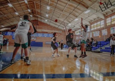 A photograph of an active WSBC basketball game in the ASDB gymnasium. On court are the competing Idaho Raptor and Washington Terrier boys teams. In the photo, a leaping Terrier player lobs a pass over the waving arms of a defender.