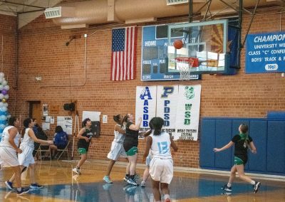 A photograph of an active WSBC basketball game in the ASDB gymnasium. On court are the competing Washington Terrier and Phoenix Roadrunner girls teams. In the photo, both teams eagerly watch as a shot attempt flies towards basket.