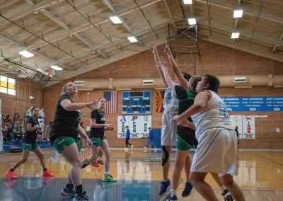 A photograph of an active WSBC basketball game in the ASDB gymnasium. On court are the competing Washington Terrier and Phoenix Roadrunner girls teams. In the photo, players from both teams watch as a Roadrunner player releases a basketball shot from the high post.