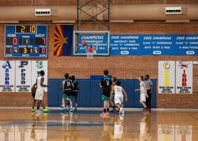 A photograph of an active WSBC basketball game in the ASDB gymnasium. On court are the competing Washington Terrier and Phoenix Roadrunner boys teams. In the photo, both teams eagerly await the basketball as it bounces off the rim of the basket