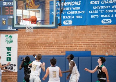 A photograph of an active WSBC basketball game in the ASDB gymnasium. On court are the competing Washington Terrier and Phoenix Roadrunner boys teams. In the photo, both teams eagerly await the basketball as it bounces off the rim of the basket.