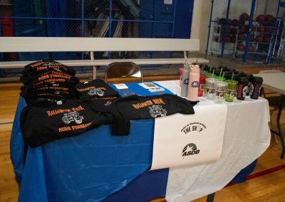 A photo of a temporary table set up in the ASDB gymnasium. Placed on top of the table are multiple black t-shirts reading, 'Halloween Bash ASDB Fundraiser'. There are also numerous cups and water bottles showing the respective logos for ASDB and WSBC. A white sign hangs from the front of the table and reads, 'Made by Students at ASDB. The Shop'.