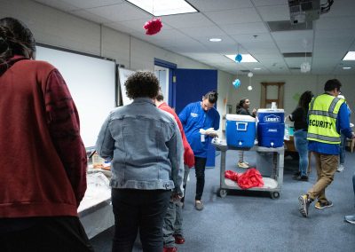 A photo of the WSBC volunteer's lounge. Multiple individuals are pictured standing in front of a temporary table in the lounge, loading up prepared food onto their plates. Two 10-gallon coolers sit to the right of the table.