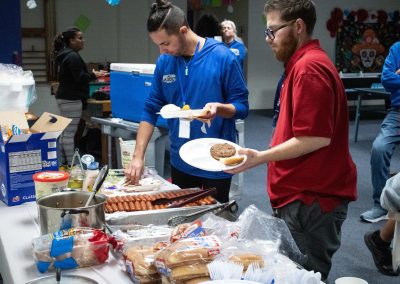 A photo of the two individuals standing in the WSBC volunteer's lounge. Before them is a large temporary table holding tins of cooked hot dogs, hamburgers, and plastic utensils. The pair are loading up the prepared food onto their plates.