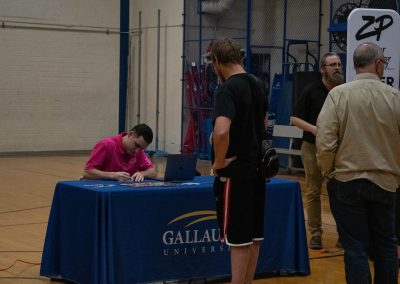 A photo of Gallaudet University's WSBC sponsorship table in the ASDB gymnasium. The table is wrapped in a royal blue table cloth and features the university's logo. A man in a pink polo shirt is seated at the table while another individual stands opposite him.