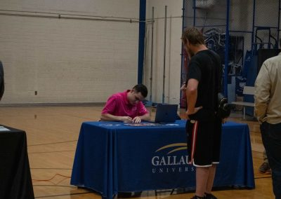 A photo of Gallaudet University's WSBC sponsorship table in the ASDB gymnasium. The table is wrapped in a royal blue table cloth and features the university's logo. A man in a pink polo shirt is seated at the table while another individual stands opposite him.