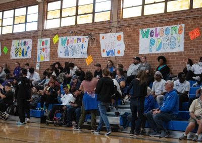 A photo of a crowd gathered in the bleachers of the ASDB gymnasium. Behind the bleachers, running the length of the gym's brick wall, are multiple posters encouraging the teams participating in WSBC.
