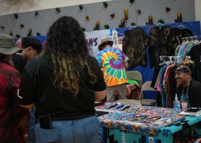 A photo of a temporary table set-up in the ASDB gymnasium. Laid out on the table are two patterned blankets, and multiple varieties of beaded necklaces. Pictured in the background are filled clothing racks.