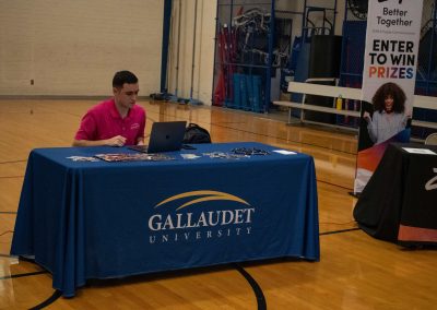A photo of Gallaudet University's WSBC sponsorship table in the ASDB gymnasium. The table is wrapped in a royal blue table cloth and features the university's logo. Sitting behind the table is a representative from the college who is wearing a pink polo shirt.