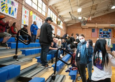 A photo of a crowd gathered in the bleachers of the ASDB gymnasium. Behind the crowd, running the length of the gymnasium's brick wall, are multiple posters encouraging the various teams participating in WSBC.