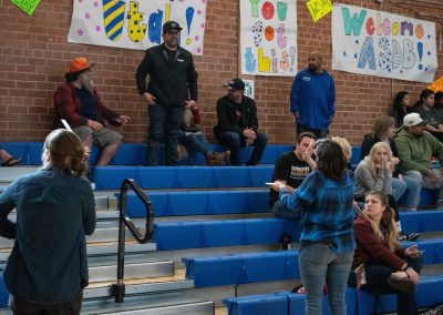 A photo of a small crowd sitting in the royal blue bleachers of the ASDB Gymnasium. Standing at the base of the bleachers are two representatives of the Tucson Deaf Community. They are signing to individuals in the crowd.