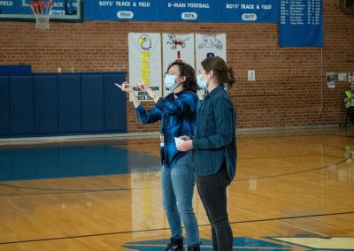A photo of two individuals in the ASDB gymnasium. They are standing at half court, signing ASL to an audience not pictured. The pair represent the Tucson Deaf Community, and are wearing blue jeans and long sleeve, button-up shirts.
