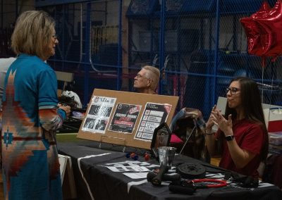 A photo of a temporary table set-up in the ASDB gymnasium. Behind the table is a young woman in a red shirt. She is signing to an older woman standing opposite her. The table is advertising 'Fitness Craver', one of the Tucson community sponsors of WSBC.