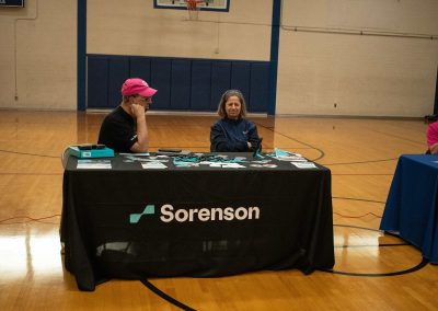 A photo of Sorenson's WSBC sponsorship table in the ASDB gymnasium. The table is wrapped in a black table cloth and features the company's logo. Two representatives from the company are pictured sitting behind the table.