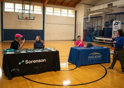 A photo of both Sorenson's and Gallaudet University's WSBC sponsorship tables in the ASDB gymnasium. The tables are covered in table cloths featuring each institutions respective logo. A pair of individuals are gathered at each table.