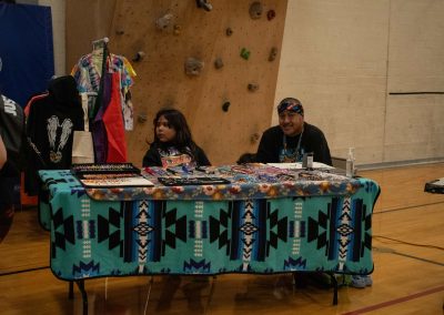 A photo of a temporary table set up in the ASDB gymnasium. Laid out on the table are two patterned blankets, as well as multiple varieties of beaded necklaces and earrings. Sitting behind the table are two individuals, one of whom is smiling for the photo.