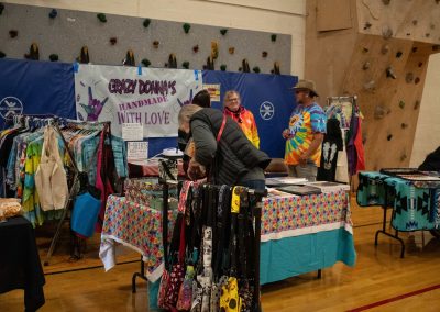 A photo of a temporary table set-up in the ASDB gymnasium. The table is topped with multiple binders, and is surrounded by racks of t-shirts and patterned handbags. Hung up behind the table is a large sign reading, 'Crazy Donna's Handmade With Love'.