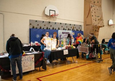 A photo of multiple temporary tables set up in the ASDB gymnasium. The tables are arranged in a straight line, and each is occupied by a different WSBC community sponsor. In the foreground, two individuals standing at the 'Jr. Sour Bar' table pose for the photo.