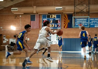 A photograph of an active WSBC basketball game in the ASDB gymnasium. On court are the competing Utah Eagle and Phoenix Roadrunner boys teams. In the photo, a Roadrunner player strides across half court with an Eagle defender trailing just behind.