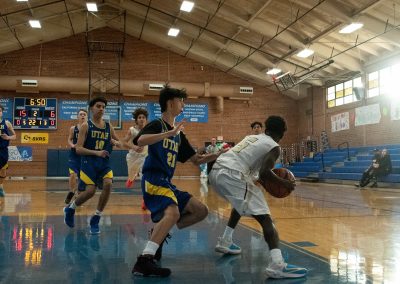 A photograph of an active WSBC basketball game in the ASDB gymnasium. On court are the competing Utah Eagle and Phoenix Roadrunner boys teams. In the photo, a Roadrunner player looks for an open teammate while being closely guarded by a defender.