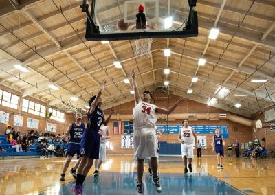 A photograph of an active WSBC basketball game in the ASDB gymnasium. On court are the competing Oregon Panther and Idaho Raptor boys teams. In the photo, a Panther player attempts a layup over the arm of a Raptor defender.
