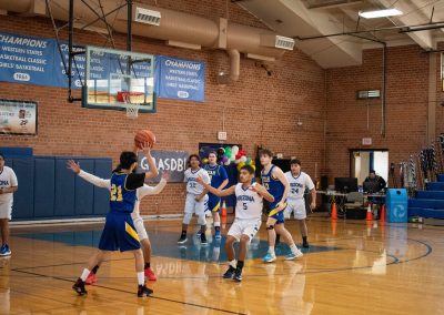 A photograph of an active WSBC basketball game in the ASDB gymnasium. On court are the competing Utah Eagle and Arizona Sentinel boys teams. In the photo, an Eagle player looks to perform an overhead pass to an open teammate.