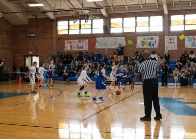 A photograph of an active WSBC basketball game in the ASDB gymnasium. On court are the competing Utah Eagle and Arizona Sentinel girls teams. In the photo, a loose basketball bounces across the court towards an Eagle player.