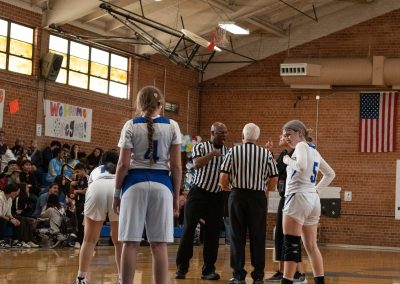A photograph of an active WSBC basketball game in the ASDB gymnasium. On court are the competing Utah Eagle and Arizona Sentinel girls teams. In the photo, the game's referees are picturing deliberating with one another.
