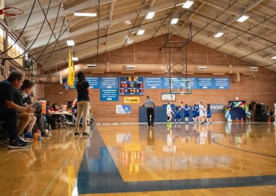 A photograph of an active WSBC basketball game in the ASDB gymnasium. On court are the competing Utah Eagle and Arizona Sentinel girls teams. In the photo, the Eagles' Head Coach watches as her team attempts to score on the opposite end of the court.