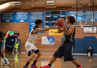 A photograph of an active WSBC basketball game in the ASDB gymnasium. On court are the competing Phoenix Roadrunner and Oregon Panther boys teams. In the photo, a Roadrunner player looks for an offensive move while being guarded by a determined Panther defender.