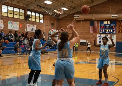 A photograph of an active WSBC basketball game in the ASDB gymnasium. On court are the competing Phoenix Roadrunner and Oregon Panther girls teams. In the photo, a determined Roadrunner player prepares to receive a lob pass from a teammate.