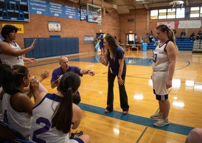A photograph of an active WSBC basketball game in the ASDB gymnasium. On court are the competing Phoenix Roadrunner and Oregon Panther girls teams. In the photo, the Oregon coaching staff are shown signing with their players during a timeout break.