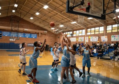A photograph of an active WSBC basketball game in the ASDB gymnasium. On court are the competing Phoenix Roadrunner and Oregon Panther girls teams. In the photo, a Roadrunner player prepares to collect a rebounding basketball with both hands.
