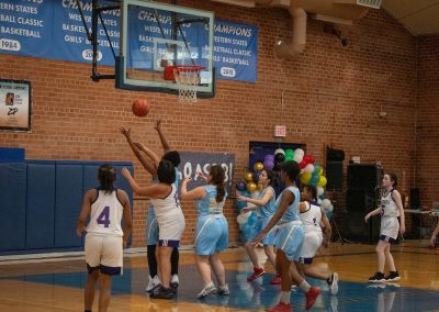 A photograph of an active WSBC basketball game in the ASDB gymnasium. On court are the competing Phoenix Roadrunner and Oregon Panther girls teams. In the photo, a Roadrunner player prepares to collect a rebounding basketball just beyond the reach of an Oregon defender.