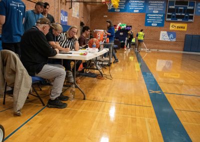 A photo of the WSBC scorekeeper's table inside the ASDB gymnasium. Seated at the table are three individuals, one of whom is writing on a piece of paper.