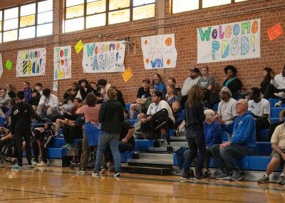 A photograph of a large crowd watching an active WSBC basketball game in the ASDB gymnasium. Behind the crowd, running the length of the gym's brick wall, are multiple posters encouraging the teams participating in WSBC.
