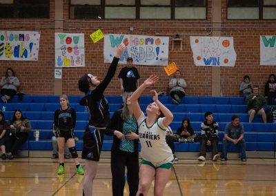A photo of ASDB Superintendent Annette Reichman leading the basketball tip off. She is flanked on either side by a respective player from the Arizona Sentinel and Washington Terrier girls teams. In the photo, both players leap for the basketball in pursuit of the rebound.