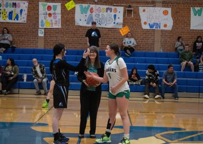 A photo of ASDB Superintendent Annette Reichman preparing to lead the basketball tip off. She is flanked on either side by a respective player from the Arizona Sentinel and Washington Terrier girls teams. Each player is in position to jump for the basketball.
