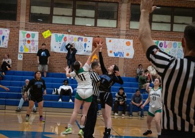 A photograph of an active WSBC basketball game in the ASDB gymnasium. Standing at half-court is a respective player from both the Arizona Sentinel and Washington Terrier girls teams. In the photo, both are leaping to rebound the basketball directly after tipoff.