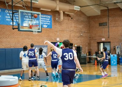 A photograph of an active WSBC basketball game in the ASDB gymnasium. On court are the competing Oregon Panther and Arizona Sentinel boys teams. In the photo, a Panther player has just attempted a three-point shot with hopes of a swish.
