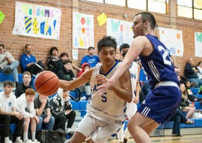 A photograph of an active WSBC basketball game in the ASDB gymnasium. On court are the competing Oregon Panther and Arizona Sentinel boys teams. In the photo, a Sentinel player is driving past a defender towards the basket.
