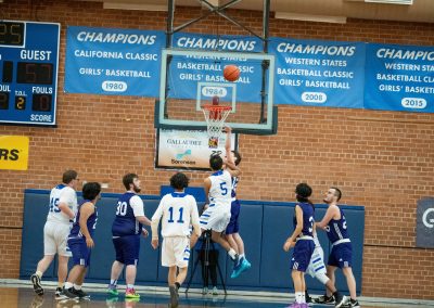 A photograph of an active WSBC basketball game in the ASDB gymnasium. On court are the competing Oregon Panther and Arizona Sentinel boys teams. In the photo, a Panther player is attempting a lay-up over the head of a leaping Sentinel defender.